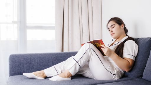 A Caucasian female in white pajamas sitting on a couch and working on her laptop
