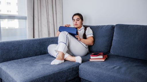 A Caucasian female in white pajamas sitting on a couch and working on her laptop