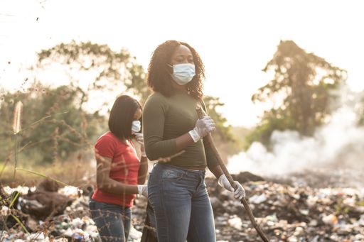 A Nigerian women cleaning up together a dump site with trees and smoke in the background in Nigeria