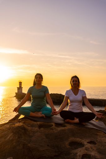 A vertical shot of two Spanish women doing yoga exercises on a coastal rock at sunset