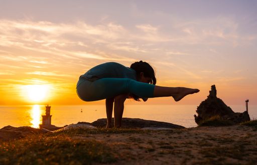 A flexible Hispanic woman doing meditation and yoga exercises on a rock at sunset next to a lighthouse