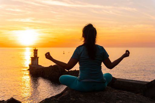 A rear view of a woman doing meditation on a rock at sunset next to a lighthouse in the sea