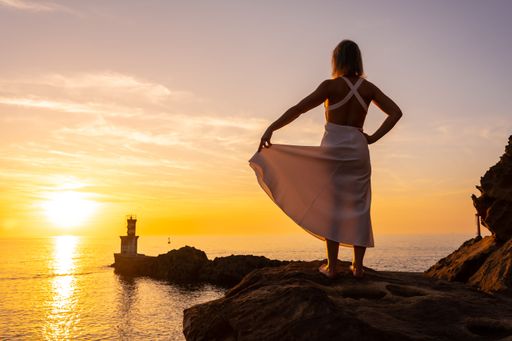 A rear view of a blonde woman in a white dress posing at sunset next to a lighthouse in the sea