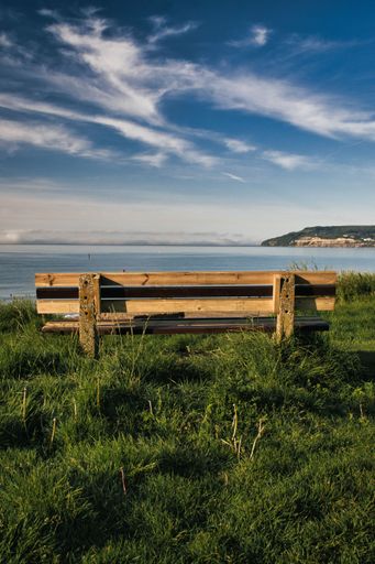 A vertical back shot of a wooden bench with a seascape under a blue cloudy sky