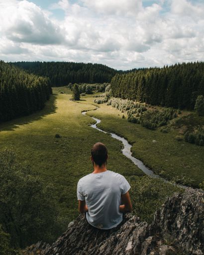 A back view of male sitting on a rock and looking at the forest with a river and large trees