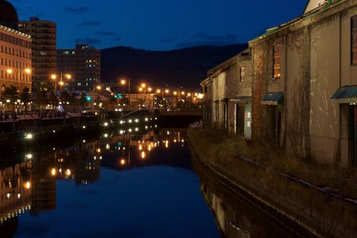 A wide shot of grass and buildings by a canal in the city of Otary-Shi in Japan during nighttime