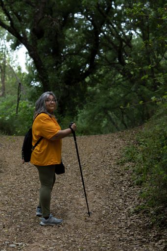 white-haired woman with walking stick, backpack and orange T-shirt hiking on a hiking trail