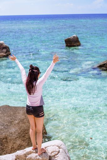 A young woman standing on a giant rock in front of the crystal clear sea with her hands up