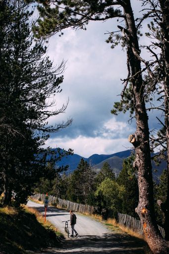 A vertical shot of a person with a bicycle on the road surrounded by hills under a cloudy sky