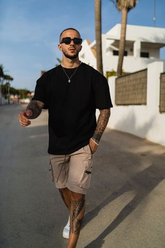 A vertical shot of a young tattooed man wearing a black t-shirt and beige shorts walking on the street