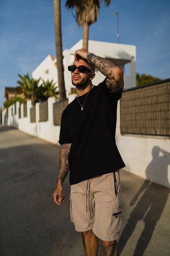 A vertical shot of a young tattooed man wearing a black t-shirt and beige shorts on the street
