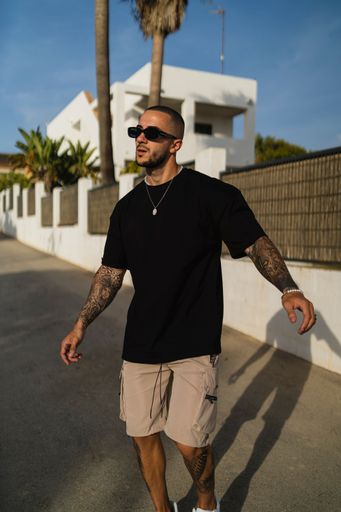 A vertical shot of a young tattooed man wearing a black t-shirt and beige shorts