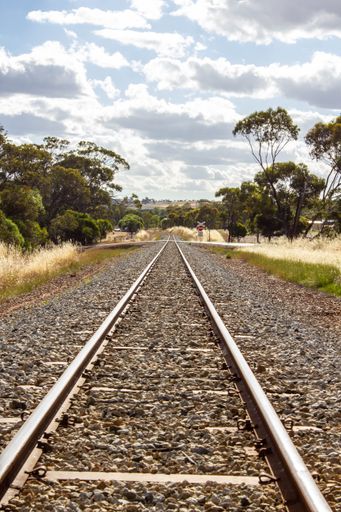 A vertical shot of train tracks through the field with green trees on blue cloudy sky background