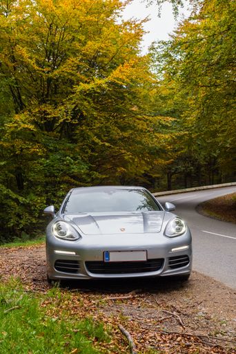 A silver Porsche Panamera S4 luxury car parked next to a winding road in a forest with autumn foliage