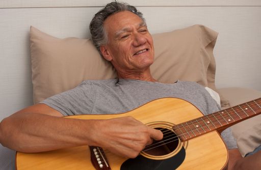 Happy Mature Man lying in bed playing his acoustic guitar.