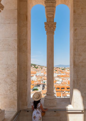 A vertical of a Caucasian female in a dress and hat looking out of a cathedral tower in Split, Croatia