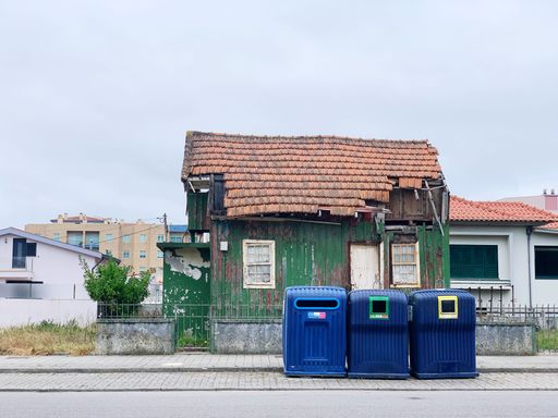 A row of garbage bins on a sidewalk with an old ruined house in the background