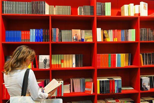 A back view of a woman reading a book at a bookshop in front of red bookshelf