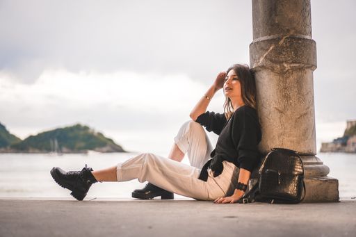 A shallow focus of a young female with long hair sitting on the ground of a building and leaning on a column surrounded by the sea