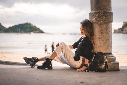 A shallow focus of a young female with long hair sitting on the ground of a building and leaning on a column surrounded by the sea