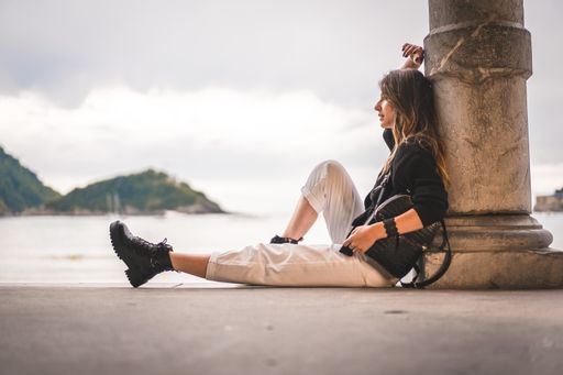 A shallow focus of a young female with long hair sitting on the ground of a building and leaning on a column surrounded by the sea