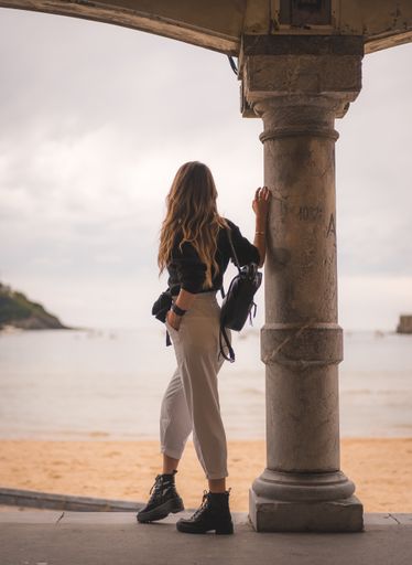 A shallow focus of a young female with long hair leaning on a column of a building surrounded by the sea