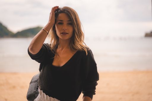 A shallow focus of a young attractive female in a black long sleeve shirt touching her hair with the sea on the background