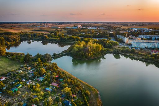 A drone shot of a beautiful lake surrounded by a green landscape with small houses under cloudy sky with sunset