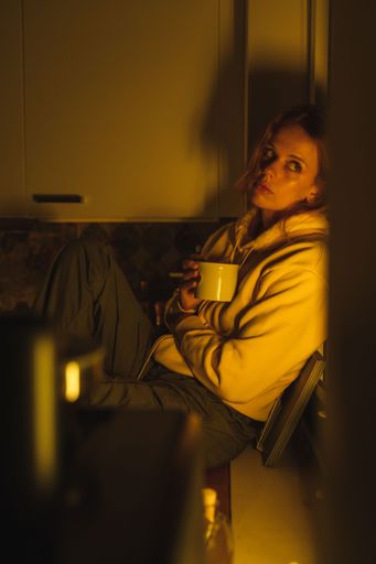 A vertical shot of a blonde girl with a mug sitting near window in yellow low-key lighting