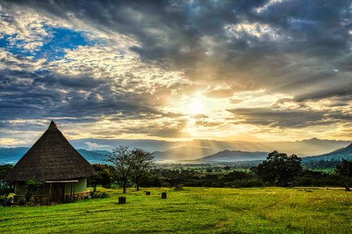 A dramatic sunrise view with sun rays shining through the clouds, a cozy hut, picturesque trees, and mountains in the background, Africa