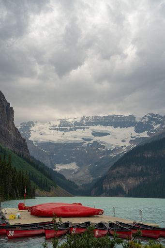 A vertical shot of the turquoise Lake Louise with rental canoes surrounded by high mountains in clouds
