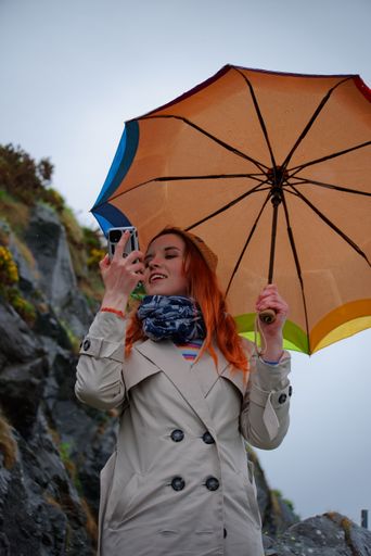 Vertical shot of beautiful young woman under colorful umbrella taking pictures with smartphone on hiking trail in foggy and rainy weather.