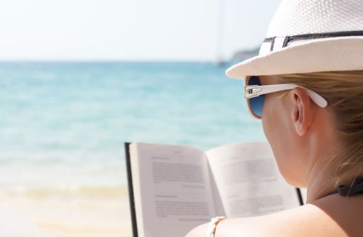 A shallow focus shot of a female with sunglasses reading a book on the beach