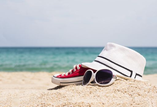 A selective focus shot of sunglasses, a white hat and red sneakers on a sandy beach