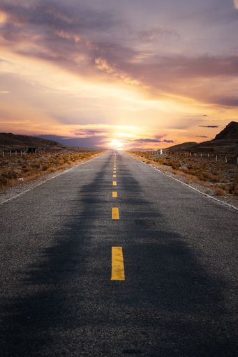 A vertical shot of a sunny asphalt road with desert sand on the sides and an orange sunset sky on the horizon