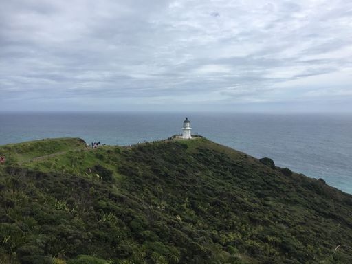 The Cape Reinga Lighthouse in New Zealand on top of a hill with a view of the sea under a bright cloudy sky