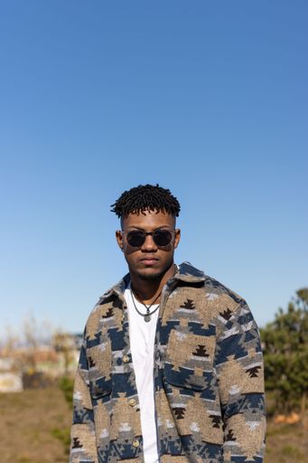 A vertical shot of a young Hispanic male with short Afro hair and sunglasses posing outside under a blue sky