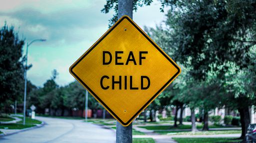 A yellow "Deaf Child" sign is posted onto a light pole on an empty neighborhood street with trees, houses, and an open street behind it