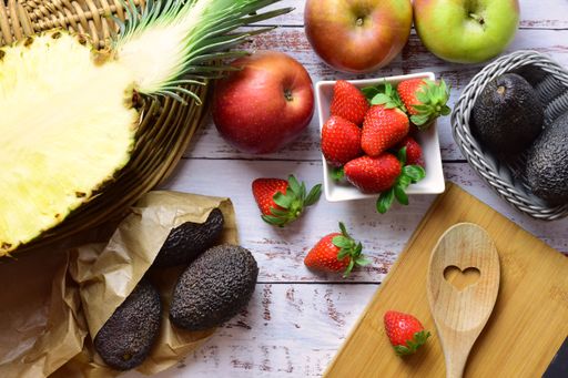 A flat lay of varied fresh fruit with strawberries, apples, pineapple and avocados on rustic white wood