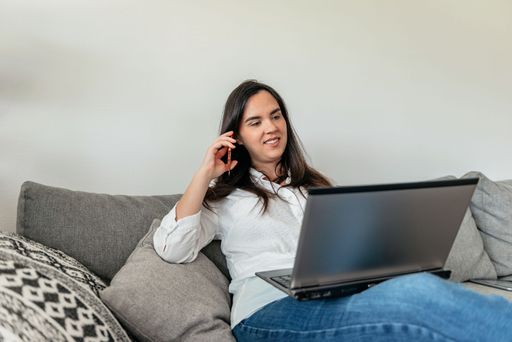 A young smiling woman sitting on a couch and talking on the phone with a laptop on her knees