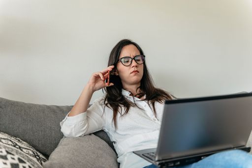 A closeup of a young displeased woman in glasses talking on the phone on a sofa looking at a laptop