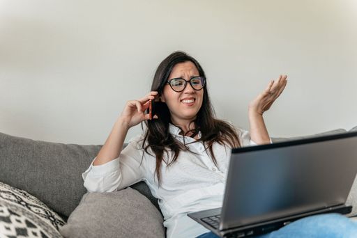 A closeup of a young perplexed woman in glasses sitting on a couch and talking on the phone with a laptop