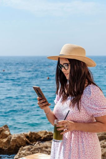A vertical shot of a woman holding her phone and enjoying a delicious drink next to a beach