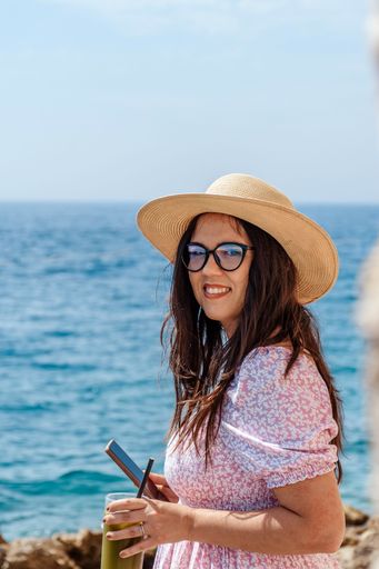 A vertical shot of a woman holding her phone and enjoying a delicious drink next to a beach