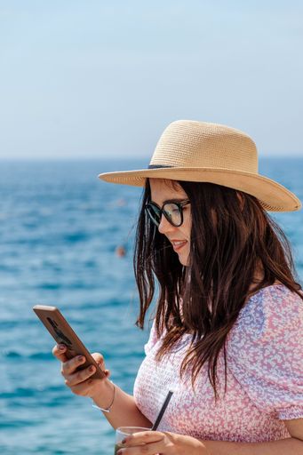 A vertical shot of a white Caucasian young girl looking at her phone in a blurred sea background
