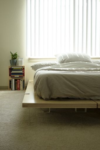A vertical interior shot of a bedroom with the mattress near a window and the bookshelf