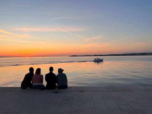 People sitting at a shore and watching a beautiful sunset in Zadar, shot from the back