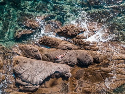 An aerial top view of a young female lying on a rocky cliff  near the wavy sea at the coasts of Greece