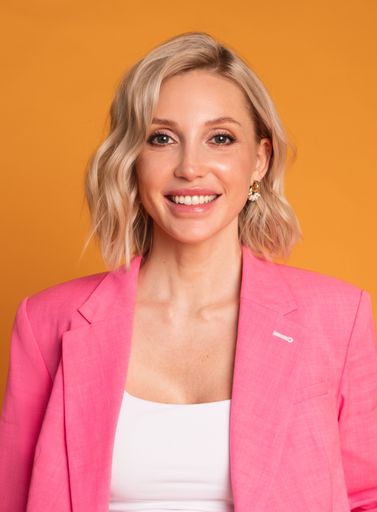 A smiling blonde Russian woman in a pink casual suit against a yellow background