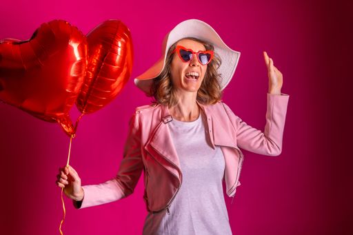 A happy woman in white hat with heart-shaped balloons ,enjoying dancing in a nightclub, on a pink wall background